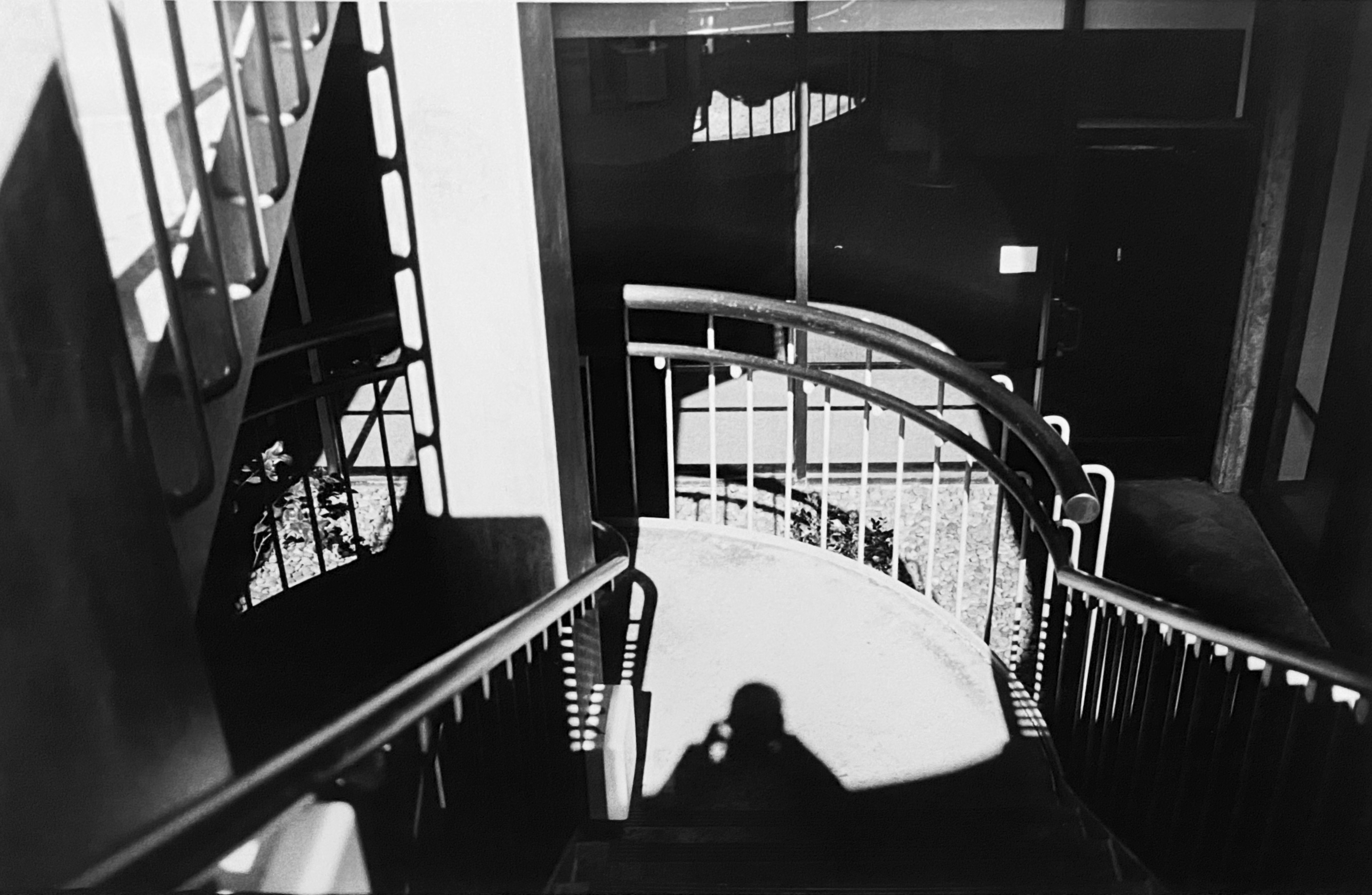 A black-and-white photograph of a spiral staircase at noon, where strong sunlight creates dramatic contrasts of light and shadow. The stair railings are half illuminated and half in darkness, while the shadows of the fence and the photographer are cast across the staircase floor.